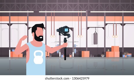 man athlete fitness blogger shooting selfie video guy in front of DSRL camera recording himself using motorized gimbal stabilizer social media concept modern gym interior horizontal