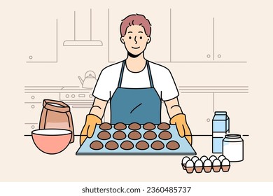 Man baker prepares oatmeal cookies or gingerbread while standing near kitchen table with ingredients. Baker guy dressed in apron and gloves proudly demonstrates dessert ready for baking.