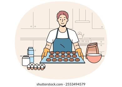 Man baker prepares oatmeal cookies or gingerbread while standing near kitchen table with ingredients. Baker guy dressed in apron and gloves proudly demonstrates dessert ready for baking.