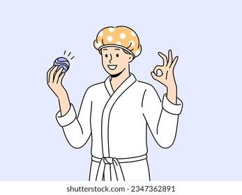 El hombre con bomba de baño está vestido con albornoz y vendaje para proteger el pelo de la humedad. Un chico joven muestra un gesto correcto recomendando el uso de una bomba de baño aromática mientras realiza procedimientos de higiene.