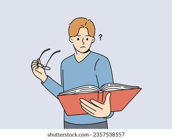 Man with book takes off glasses, trying to make out text in textbook and getting upset because of deterioration of vision. Guy is experiencing vision problems associated with excessive eye strain.