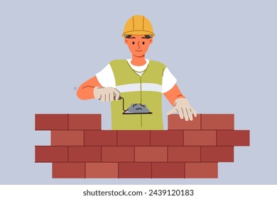 Man bricklayer builder builds brick wall using trowel with concrete mixture to secure blocks. Guy bricklayer in yellow vest and hardhat works in architectural company performing contract work