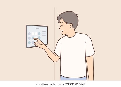 Man controls smart home system using digital panel hanging on wall to turn iot devices on and off. Guy is standing in room adjusting temperature and lighting in apartment equipped with smart home