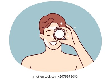 Der Mann zeigt ein Glas Sahne, das im Kosmetikgeschäft gekauft wurde, um sein Gesicht zu befeuchten oder Falten loszuwerden. Junger Mann mit nackten Schultern empfiehlt, nährenden Balsam oder Creme für die Hautpflege zu kaufen