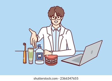 Man dentist pulls hand to screen, sitting at table with laptop and supplies for dental care. Young doctor dentist recommends purchasing toothbrush and toothpaste to maintain hygiene