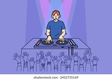 Man DJ performs in nightclub spinning records on mixing console, near crowd of partygoers with hands raised. Cheerful guy DJ in headphones plays dance hits for party or festival visitors