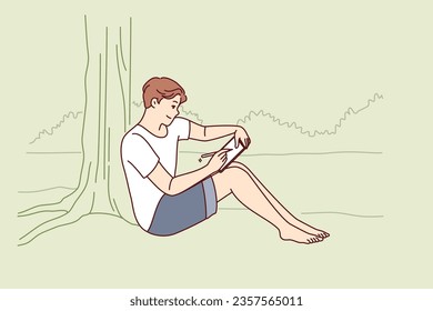 Man draws in electronic tablet sitting on shore of lake and enjoying beautiful landscape of morning nature. Creative young man is experiencing surge of inspiration being in park with river