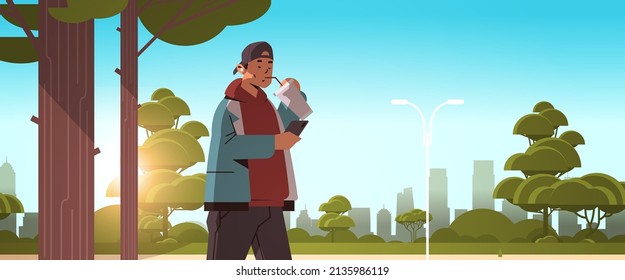 man drinking coffee with straw and using smartphone while walking in urban park social media communication digital addiction