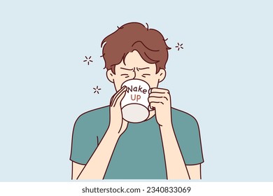 Man drinks coffee from mug with inscription wake up, wanting to cheer up and gain strength before difficult working day. Tired young guy trying to wake up with caffeine and taurine drinks