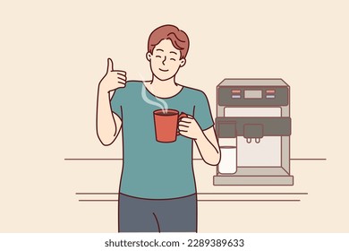 Man drinks coffee from mug standing near machine for making delicious espresso and shows thumbs up. Young guy takes coffee break to cheer up and increase productivity or stay awake doing tedious work 