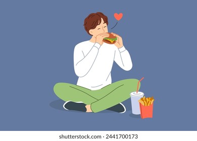 Man eats hamburger with sausage and cheese, sitting on floor and enjoying taste of fast food from street restaurant. Guy enjoys hamburger or cheeseburger prepared according to perfect recipe