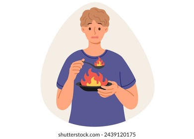 Man eats very spicy food, causing burning sensation in mouth due to overabundance of pepper, holding plate and spoon with flame. Guy eats spicy dish with spices, makes dissatisfied grimace