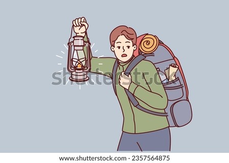 Man is engaged in hiking and carries large backpack on back, holding kerosene lantern to light way in dark. Frightened guy hears rustle in bushes and looks around while hiking in forest