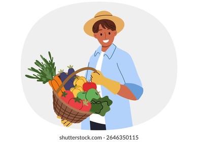 Man farmer holds basket of fresh vegetables in hands, rejoicing at excellent harvest, and smilingly looks at screen. Farmer guy in hat and gloves sells organic products filled with vitamins at fair