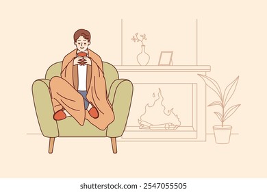 Man feels comfort and harmony sitting by fireplace with blanket on shoulders and cup of hot coffee in hands. Guy enjoys comforts of home and apartment is cozy, allowing him to relax on winter evenings