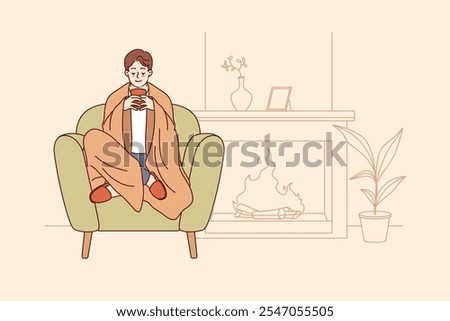 Man feels comfort and harmony sitting by fireplace with blanket on shoulders and cup of hot coffee in hands. Guy enjoys comforts of home and apartment is cozy, allowing him to relax on winter evenings