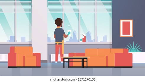 man in gloves and apron cleaning windows with rag cleaner spray rear view african american guy doing housework concept modern apartment living room interior flat full length horizontal