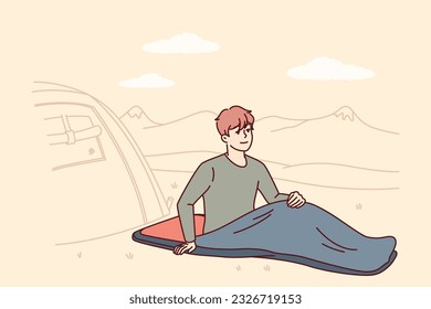 Man hiker with sleeping bag sits on ground on camping site near tent located in national natural park. Smiling guy traveler waking up in morning and spending time in camp for camping