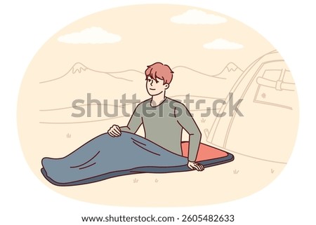 Man hiker with sleeping bag sits on ground on camping site near tent located in national natural park. Smiling guy traveler waking up in morning and spending time in camp for camping