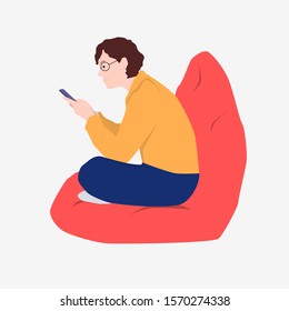 A man holding a phone sitting in a comfortable chair. A young man is reading an online book.