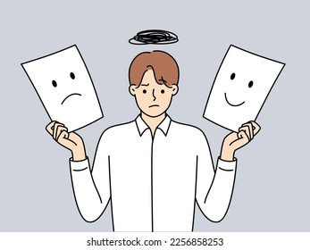 Man holds two sheets of paper with different emotions symbolizing mood swings due to work overload. Guy in shirt shows image of happy and sad mood on white sheets