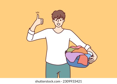 Man householder with laundry in basin shows thumbs up recommending quality laundry detergent. Guy getting ready to load textiles into washing machine setting positive example for peers