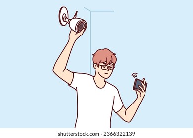 Man installs surveillance camera on wall and holds mobile phone, watching video through WIFI network. Guy establishes video surveillance system, wanting to secure home or office from criminals.