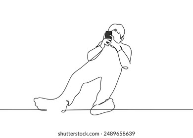 man lies at full height with his feet towards the camera holding a phone with both hands - one line art vector. concept mobile surfing, social media addiction. Handmade vector not AI