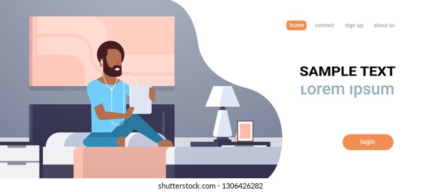 man listening audio book through headphones african americn guy using tablet sitting on bed modern home bedroom interior male character full length flat horizontal copy space