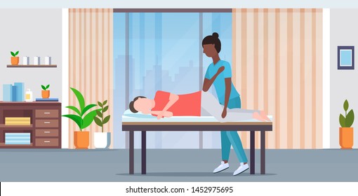 man lying on massage bed african american masseuse therapist doing healing treatment massaging injured patient manual physical therapy concept modern hospital office interior horizontal full length