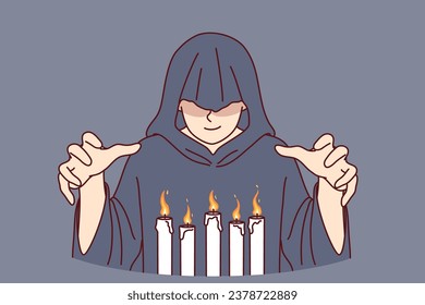 Man monk in hood stands over burning candles, performing mysterious religious ritual to communicate with otherworldly world. Monk experiments with black magic and practices witchcraft