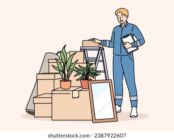 Man mover stands near pile of cardboard boxes advertising services of company that helps with moving to new home. Mover guy in blue uniform works in logistics business providing delivery services.