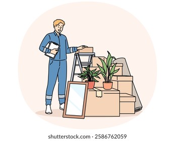 Man mover stands near pile of cardboard boxes advertising services of company that helps with moving to new home. Mover guy in blue uniform works in logistics business providing delivery services.