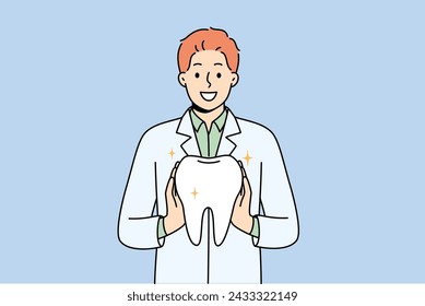 Man orthodontist from dental clinic holds model of healthy tooth, offering you procedure to get rid of caries. Guy orthodontist in white coat smiles, recommending becoming client of new dental center