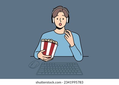 Mann mit Paket Popcorn sieht Film sitzend am Tisch mit Computer und sieht auf Bildschirm fasziniert. Funny in Kopfhörern ist entspannt beim Anschauen neuer Filmblockbuster oder beliebter Fernsehserie