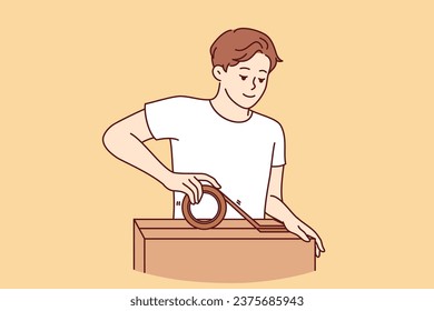 Man packs personal belongings into cardboard relocation box and seals lid with tape. Positive guy small business owner holding box and preparing goods ordered by customer for shipment