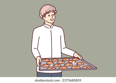 Man pastry chef is holding tray of cookies, dressed in white chef shirt and disposable hair cap. Guy confectioner prepares oatmeal cookies from natural ingredients, for sale in own bakery