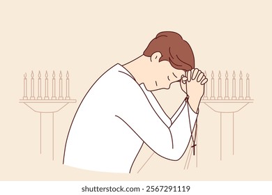 Man prays, standing in church near candlesticks, holds christian crucifix and closes eyes, leaning on crossed palm. Guy prays in example of important event, asks god for help to avoid mistakes