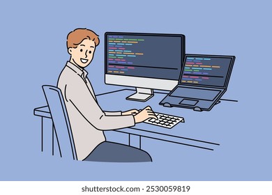 Man programmer types code and looks at camera, sitting at table with computer and laptop. Successful guy programmer develops software or application, working in team with UI or UX designers