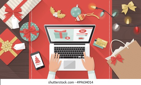 Man purchasing Christmas gifts online using a laptop on his desk, shopping bags and decorations all around, holiday and celebrations banner