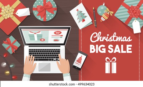 Man purchasing Christmas gifts online using a laptop on his desk, shopping bags and decorations all around, holiday and celebrations banner