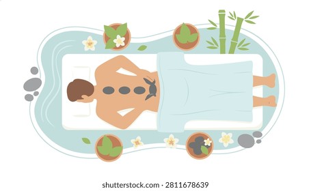 Man receiving hot stone massage treatment on spa table surrounded by bamboo and flowers in top down view.
