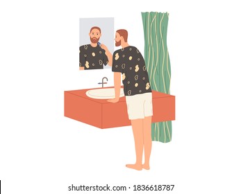 Man shaves his beard with an electric razor while standing in the bathtub by the mirror