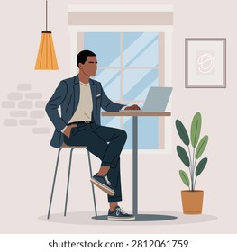 Man sits calmly at a small table with laptop. He wears a stylish blazer and comfortable sneakers. Natural light streams through the window behind him. Potted plant adds greenery beside the table