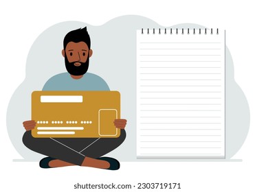 A man sits cross-legged in his hands holding a large credit card next to a close-up notepad for text. Concept of credit card, expense planning, income planning.
