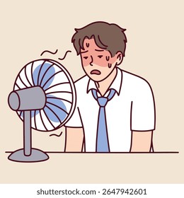 O homem fica sentado na mesa suando e limpa a testa enquanto o ventilador sopra. Ele luta com o calor e a exaustão no local de trabalho interno