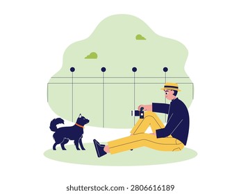Man sitting on the grass outdoors, using a small camera to photograph a dog standing near a fence.