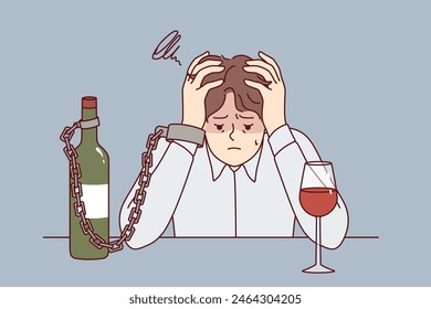 Man suffering from alcohol addiction is chained to bottle of wine, sitting at table clutching head. Guy with liquor addiction fell into depression after being betrayed by girlfriend