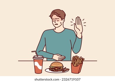 Man suffering from gastritis refuses fast food that causes pain in abdomen on advice of nutritionist. Guy near table with junk lunch shows stop gesture due to gastritis caused by eating hamburgers