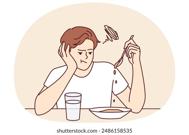 Man suffers from lack of appetite sitting at table in kitchen and does not want to eat breakfast. Depressed guy with unhappy expression melancholy looks at plate with not appetite food during diet